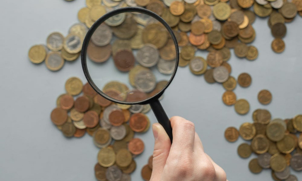 Das Bild zeigt einen auf einem Tisch ausgebreiten Haufen Münzen, die durch eine Lupe betrachtet und näher fokussiert werden.
