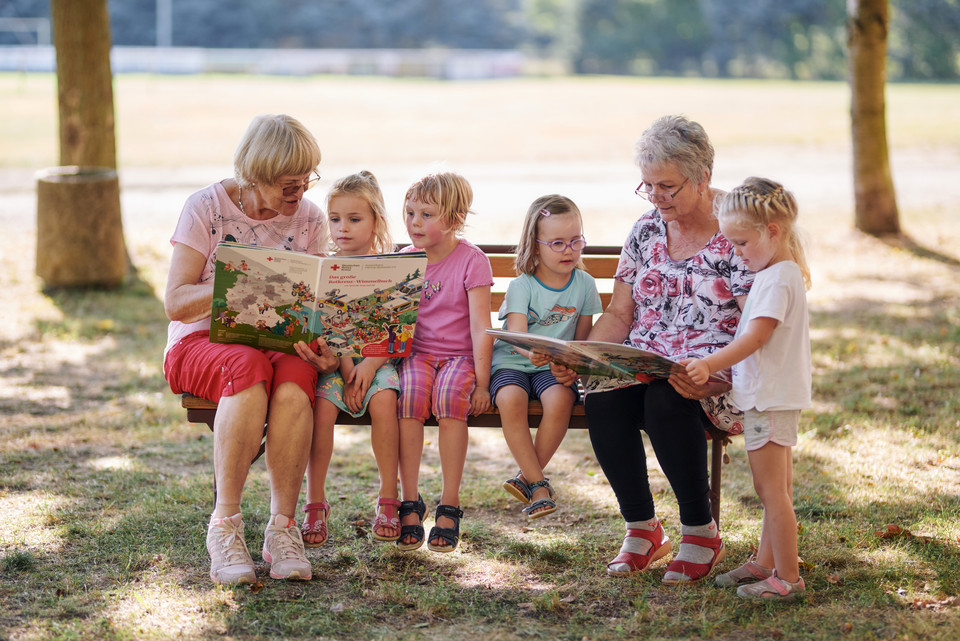 Zwei Frauen sitzen auf einer Bank im Freien und lesen mehreren Kindern aus großen Bilderbüchern vor