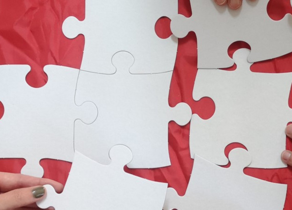 drei Hände puzzeln auf rotem Grund große weiße Puzzleteile zusammen