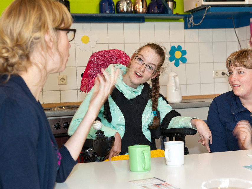 Drei Personen sitzen an einem Tisch in einer Küche. Die Person links gestikuliert mit der Hand, während die anderen beiden Personen aufmerksam zuhören. Eine Person in der Mitte sitzt in einem Rollstuhl.
