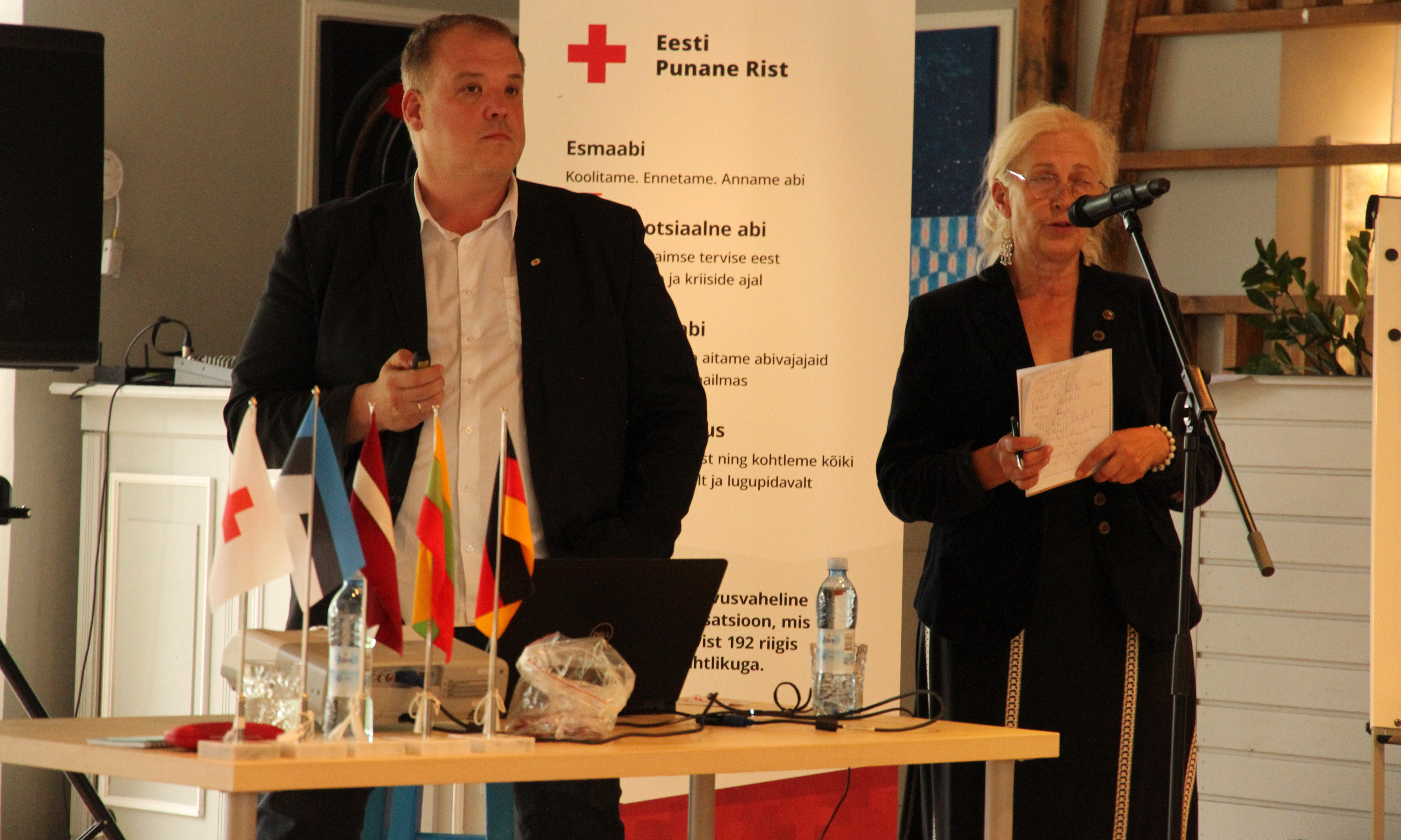 Zwei Personen sprechen in Mikrofon. Im Hintergrund ein banner mit rotem Kreuz