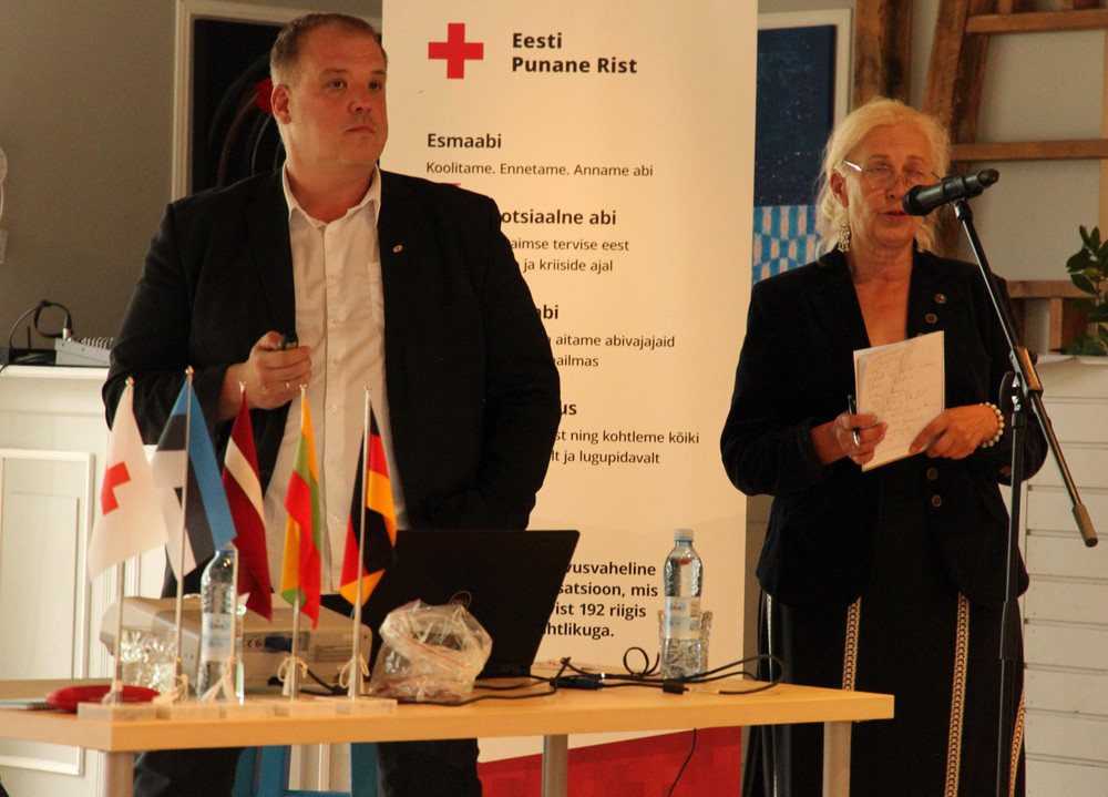 Zwei Personen sprechen in Mikrofon. Im Hintergrund ein banner mit rotem Kreuz