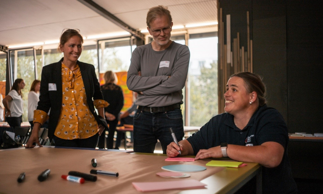 Eine Frau schreibt auf Post-its an einem Tisch, zwei Personen schauen zu. Im Hintergrund stehen Personen im Raum.