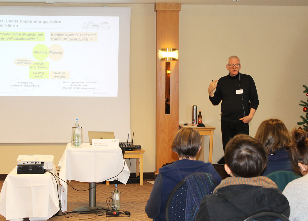Vortragender steht vor einer Leinwand mit Präsentation, Publikum sitzt auf Stühlen und hört zu, rechts ein geschmückter Weihnachtsbaum