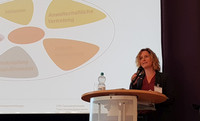 Sabine Urban steht an Stehtisch mit Mikrofon vor einer Präsentationsfolie mit fünf farbigen Segmenten in einer Ellipse.