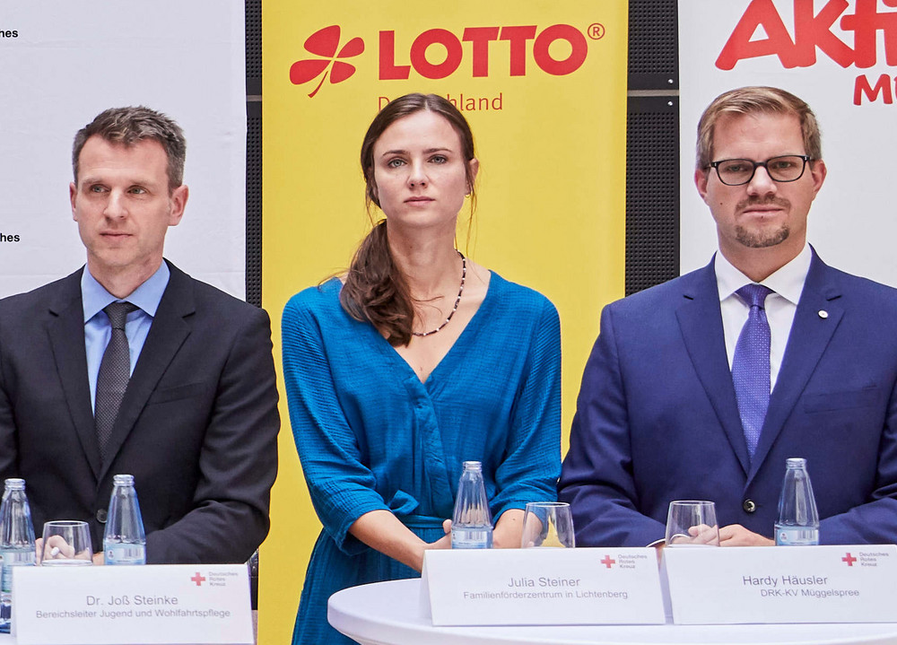 Sechs Personen stehen bei einer Pressekonferenz hinter drei Stehtischen mit weißen Hussen, im Hintergrund Banner von Deutschem Rotem Kreuz, Lotto, Aktion Mensch und Deutscher Fernsehlotterie.