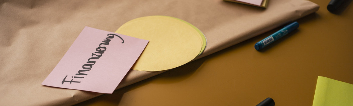Tisch mit Post-its und Stiften, auf einem Post-it steht "Finanzierung".