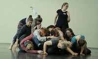 Gruppe von Menschen in Bewegung, einige liegen auf dem Boden, andere stehen oder beugen sich, in einem hellen Raum mit dunklem Boden.