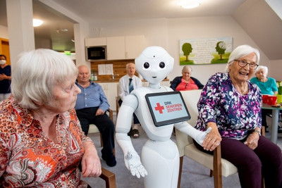Auf dem Foto ist der Roboter Pepper zu sehen, der zwischen zwei Seniorinnen sitzt. Diese Lachen und freuen sich scheinbar über den Roboter.