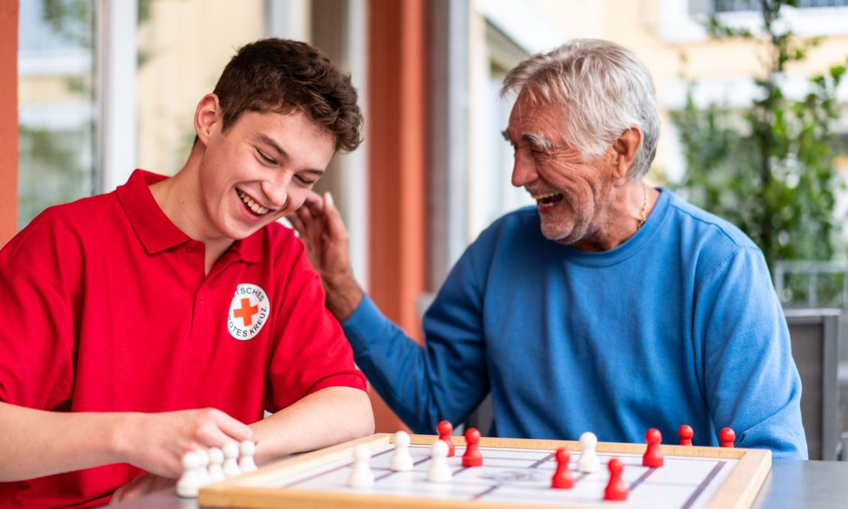 Ein junger Pfleger und ein älterer Mann sitzen am Tisch, lachend.