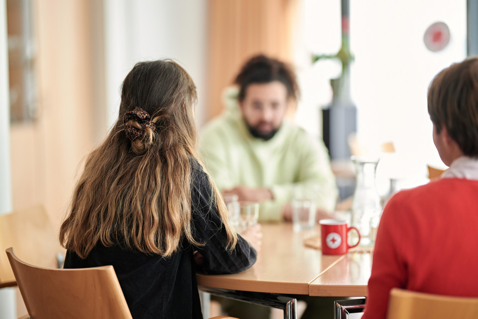Drei Personen sitzen an einem Tisch, zwei von hinten sichtbar, eine Person mit verschwommenem Gesicht im Hintergrund