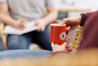 Eine Person in einer Suchtselbsthilfegruppe hält eine rote Tasse mit dem Logo des Deutschen Roten Kreuzes. Im Hintergrund sitzt eine weitere Person mit einem Notizblock.