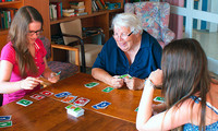 Zwei junge und eine ältere Person sitzen an einem Holztisch und spielen ein Kartenspiel mit bunten Karten in Rot, Blau, Grün und Gelb.