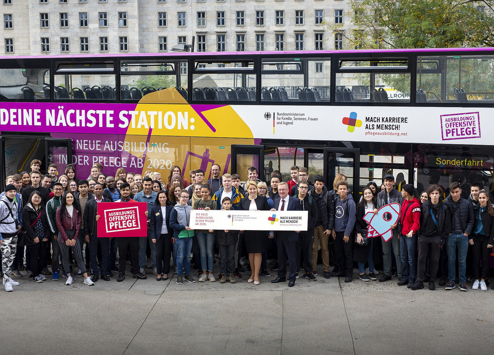 Große Gruppe vor einem zweistöckigen Bus mit Werbeaufschrift zur Pflegekampagne und mehreren Personen, die ein großes Schild halten.
