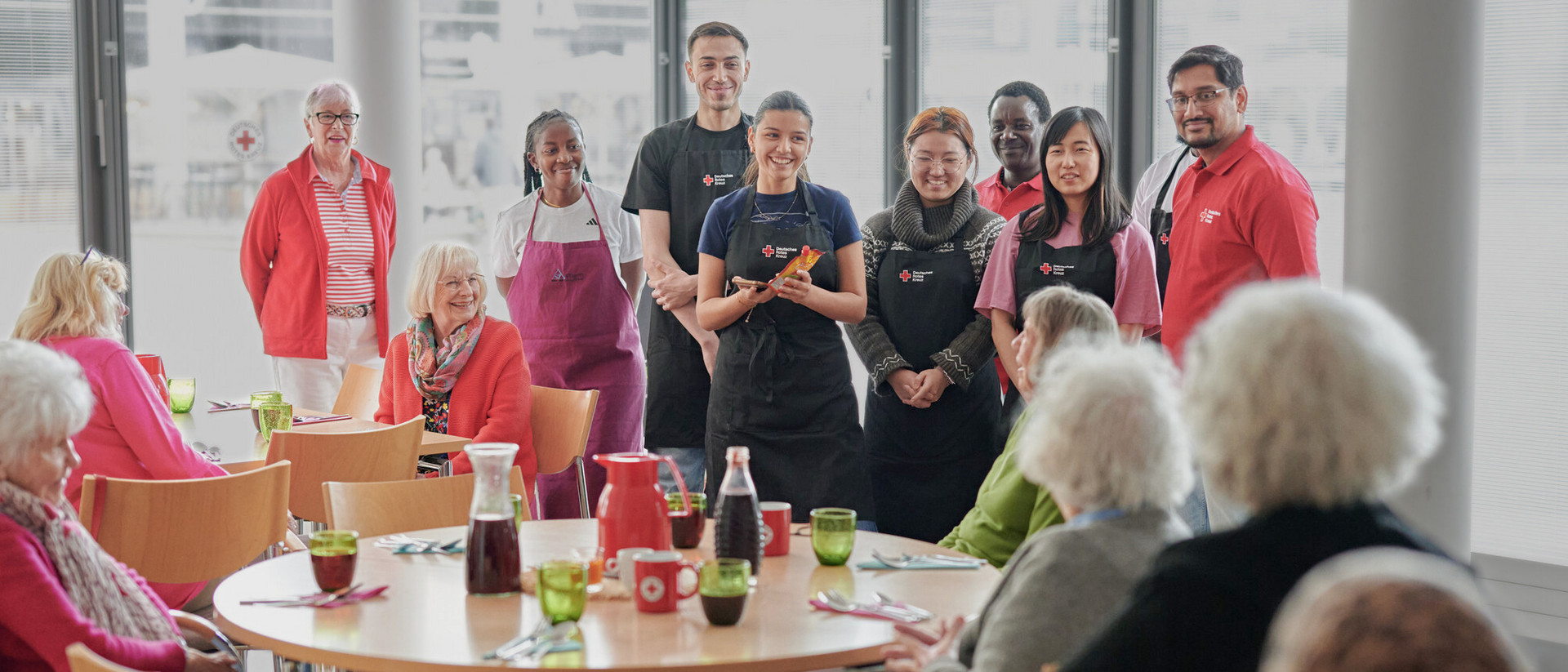 Mehrere Seniorinnen und Senioren sitzen an runden Tischen, während eine Gruppe von Menschen in Arbeitskleidung und Freizeitkleidung im Hintergrund steht.