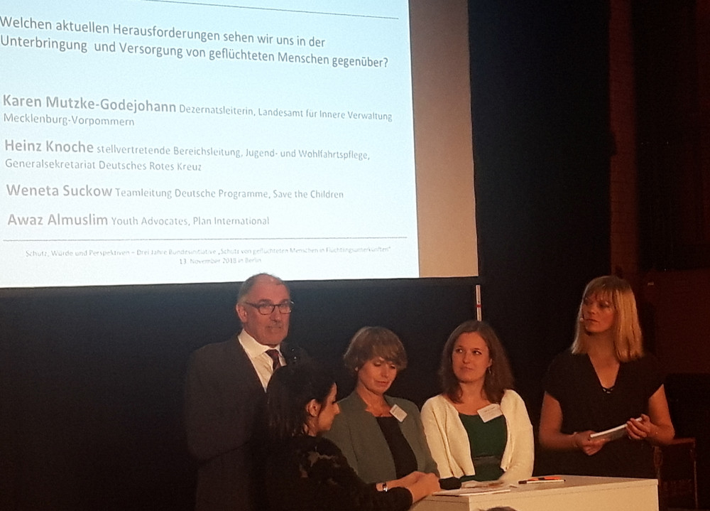 Fünf Personen stehen an einem Pult vor einer Leinwand mit Text, im Publikum sitzen mehrere Menschen in einem dunklen Raum.