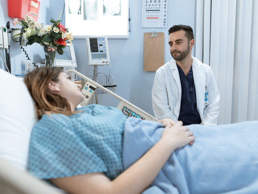 Frau liegt im Krankenhausbett und spricht mit einem Arzt im weißen Kittel im Patientenzimmer