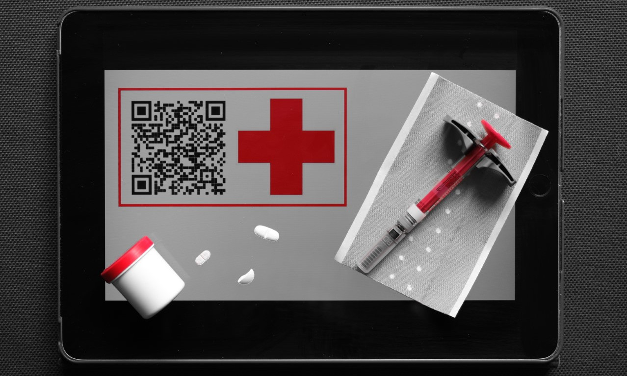 Ein DRK Tablet mit Logo, QR-Code. Darauf liegen einige Tabletten, eine Spritze und ein Pflaster.