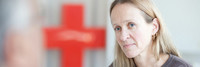 Frau schaut in Richtung der Kamera zu einem Mann, dessen Gesicht nicht erkennbar ist, im Hintergrund ist ein rotes Kreuz zu sehen