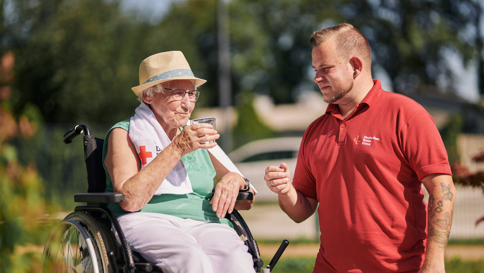 Mitarbeiter des DRK reicht bei Hitze einer Seniorin ein Glas Wasser