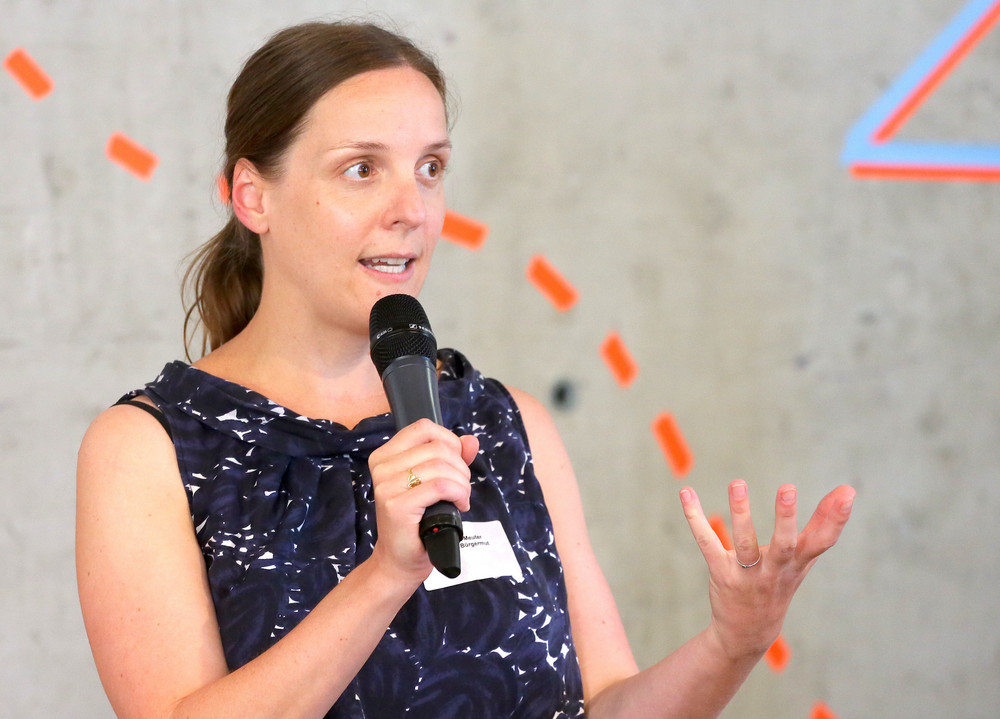 Julia Meuter hält Mikrofon und gestikuliert mit der freien Hand vor grauer Wand mit rotem Linienmuster.