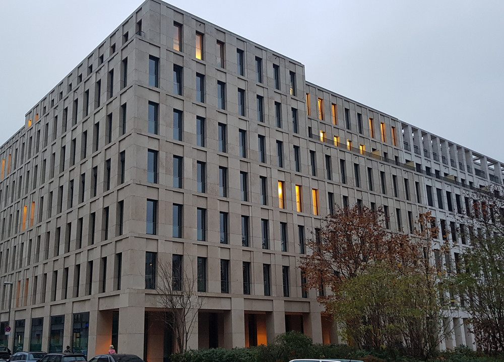 Breit aufgenommener Blick auf das mehrstöckige Gebäude des GKV mit vielen Fenstern und einigen beleuchteten Räumen, davor geparkte Autos und kahle Bäume.