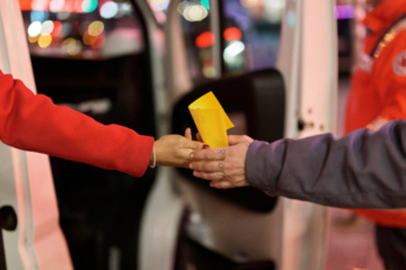 Zwei Personen reichen sich vor einem Fahrzeug einen gelben Gegenstand, eine Person trägt eine rote Jacke