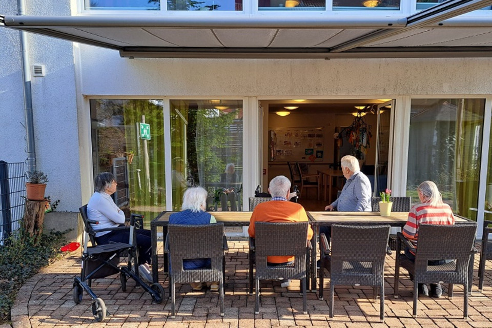 Seniorinnen und Senioren einer Wohngruppe sitzen auf einer Terrasse vor einem Wohnhaus mit Garten, erkennbar mehrere Sitzecken und Pflanzen, über ihnen eine schattenspendende Marquise
