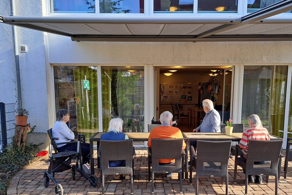 Seniorinnen und Senioren einer Wohngruppe sitzen auf einer Terrasse vor einem Wohnhaus mit Garten, erkennbar mehrere Sitzecken und Pflanzen, über ihnen eine schattenspendende Marquise