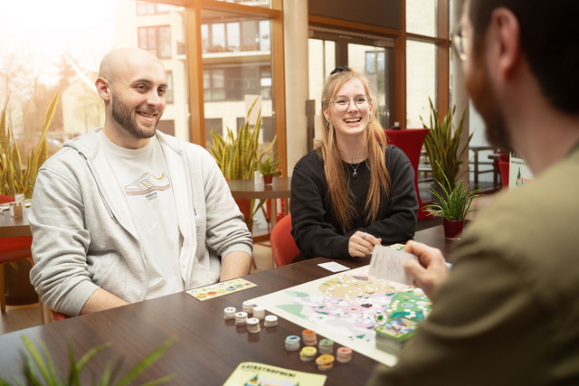 Drei Personen sitzen an einem Tisch und spielen ein Brettspiel zum Thema Katastrophenschutz mit Spielbrett, Spielkarten und Spielsteinen.
