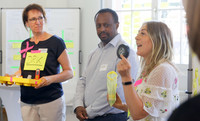 Drei Personen stehen in einem Raum, eine hält ein gelbes Tablett mit einem Modell, eine andere zeigt ein rundes Objekt, die dritte Person hält eine durchsichtige Tüte.