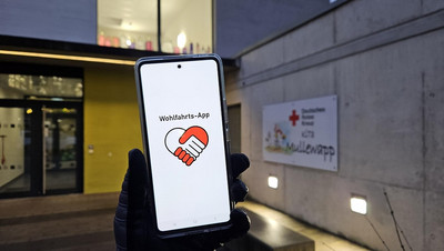 Eine Hand hält ein Smartphone hoch, auf dem das Logo der Wohlfahrts-App geöffnet ist.