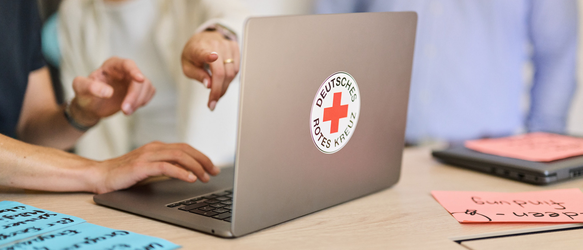 Laptop mit Aufkleber des Deutschen Roten Kreuzes auf dem Tisch, zwei Personen zeigen auf den Bildschirm