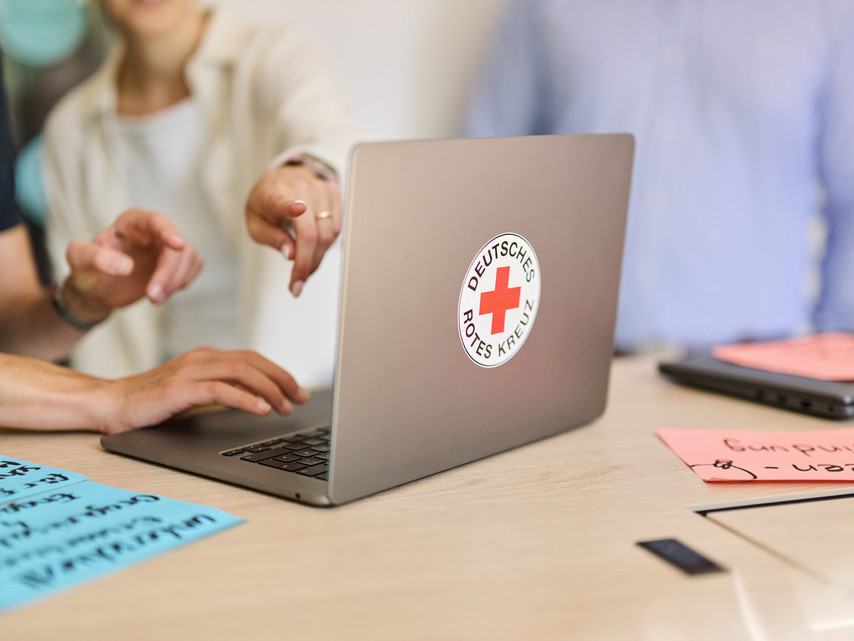 Laptop mit Aufkleber des Deutschen Roten Kreuzes auf dem Tisch, zwei Personen zeigen auf den Bildschirm