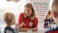 DRK-Mitarbeiterin sitzt mit Kindern am Tisch und schaut das Kind neben sich an
