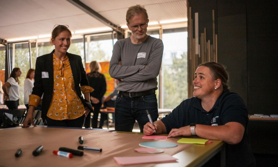 Drei Personen an einem Tisch. Die beiden Personen links im Bild stehen und schauen zur der Person rechts im Bild. Diese schaut nachlinks vorne, hält einen Stift in der Hand vor ein paar bunten Post-its und grinst.