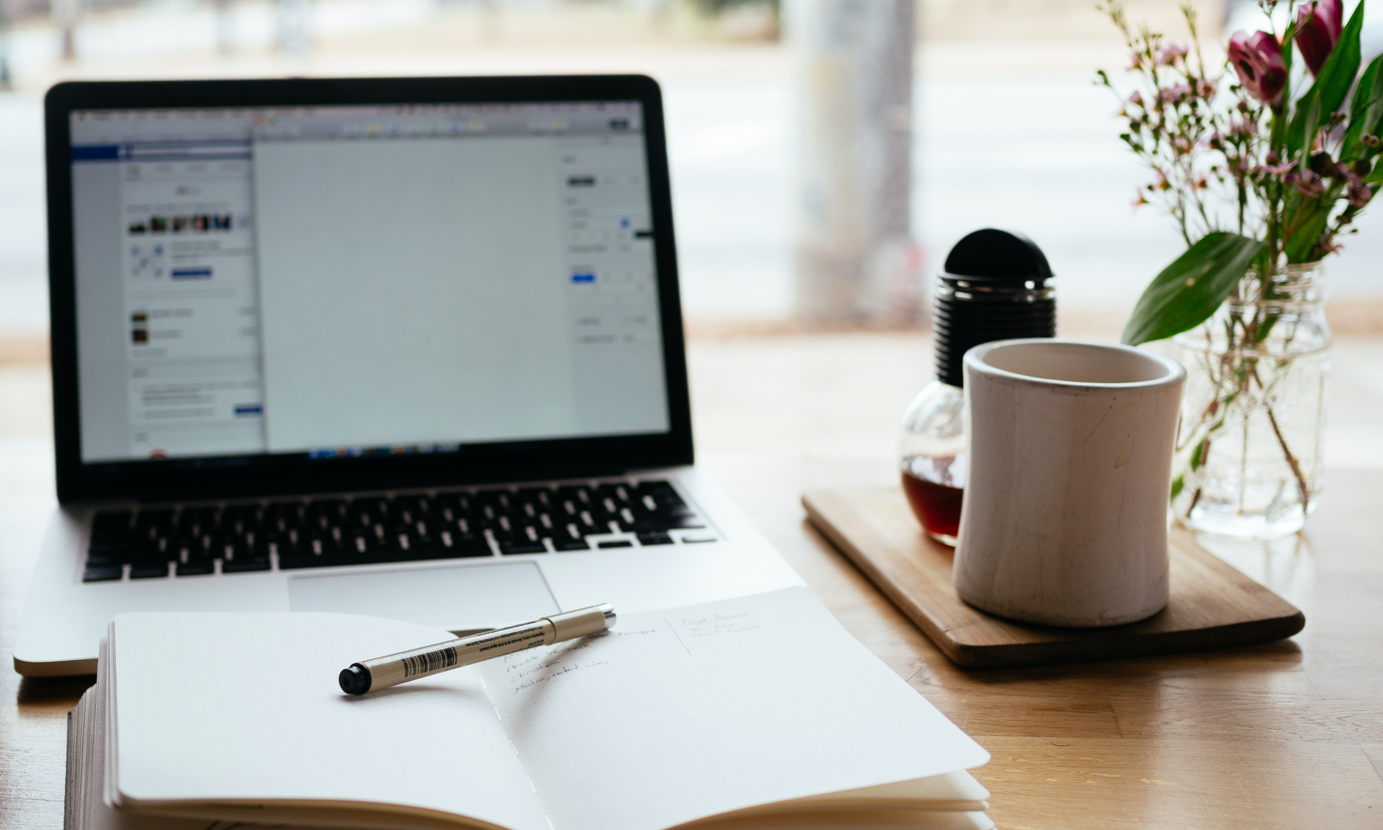 Foto eines Laptop vor dem ein Notizbuch liegt. Rechts davon eine Kaffeetasse