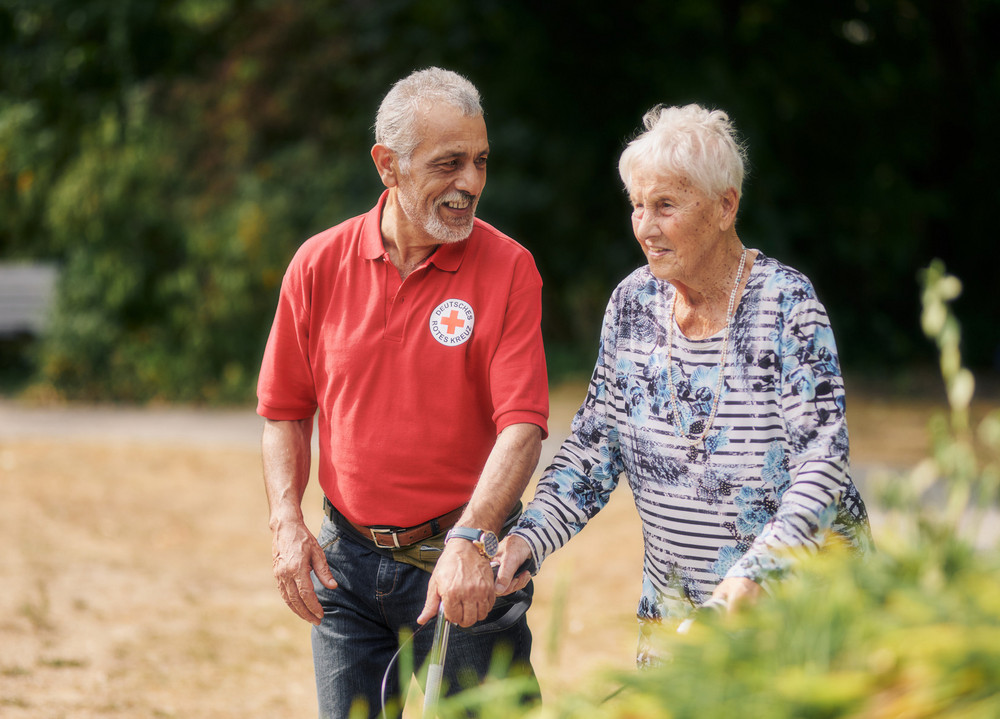 Pflegekraft mit rotem DRK Shirt spaziert mit Dame mit Gehhilfe