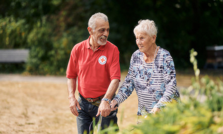 Pflegekraft mit rotem DRK Shirt spaziert mit Dame mit Gehhilfe