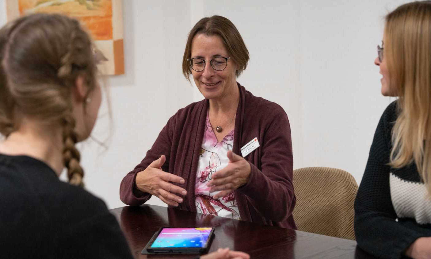 Eine Frau gestikulierend an einem Tisch vor einem Tablet und im Gespräch mit mit zwei Studentinnen.