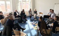 Gruppe von Menschen sitzt im Stuhlkreis in einem hellen Raum, auf dem Boden liegen viele bunte Zettel.