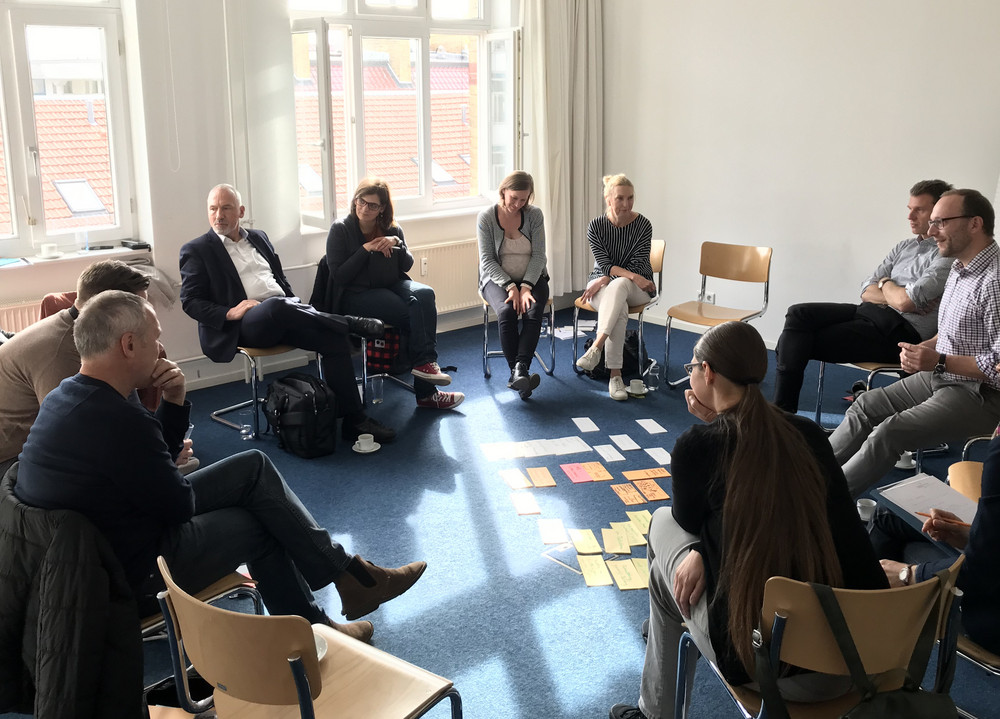 Gruppe von Menschen sitzt im Stuhlkreis in einem hellen Raum, auf dem Boden liegen viele bunte Zettel.