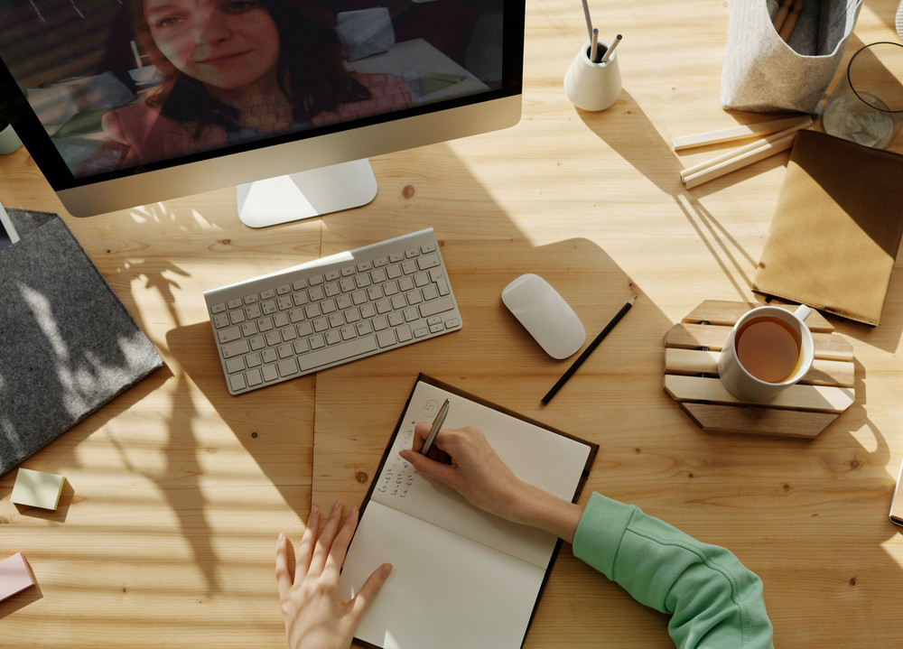 Monitor mit Videokonfernt; auf dem Tisch schreibt eine Person ins Notizbuch, weitere Büromaterialien und eine Kaffeetasse.