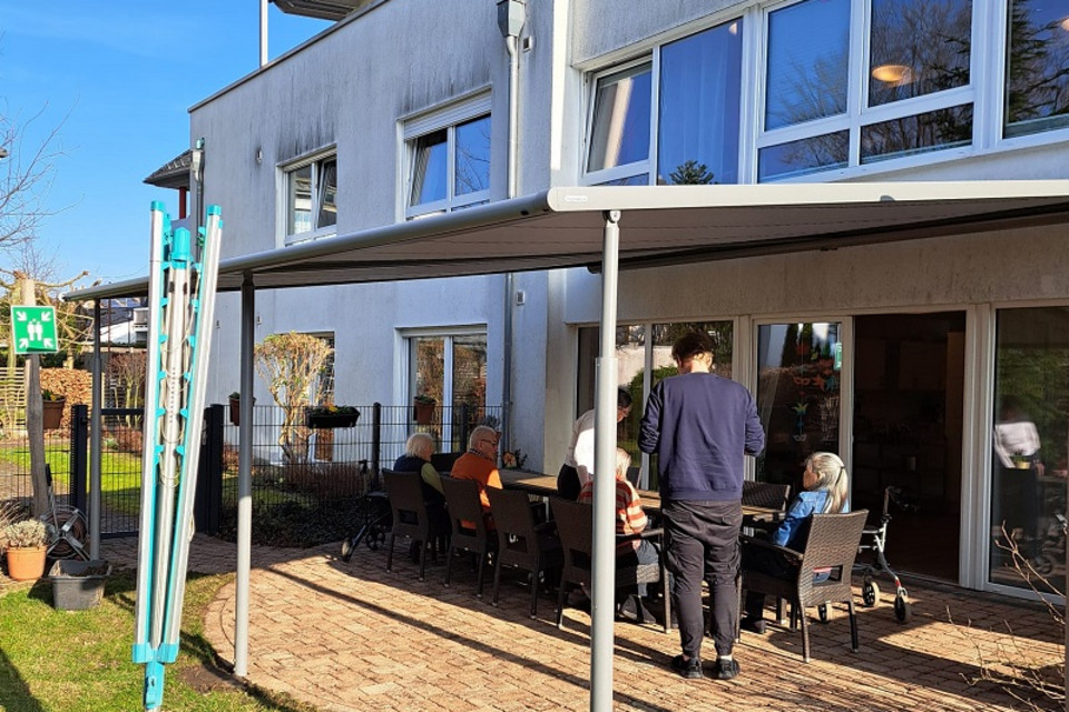 Mehrere ältere Menschen sitzen an einem langen Tisch auf einer gepflasterten Terrasse vor einem modernen Wohngebäude, ein jüngerer Mann steht daneben, im Vordergrund ein Sonnenschutz