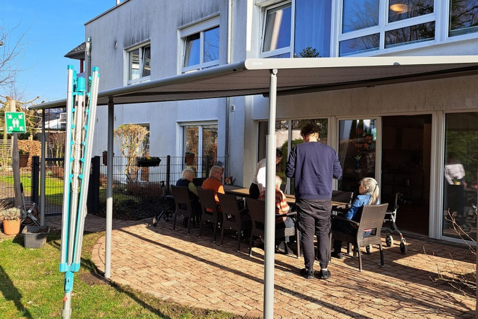 Mehrere ältere Menschen sitzen an einem langen Tisch auf einer gepflasterten Terrasse vor einem modernen Wohngebäude, ein jüngerer Mann steht daneben, im Vordergrund ein Sonnenschutz