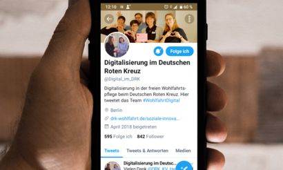 In einer Hand mit natürlichem brauen Hintergrund liegt ein Handy oder auch Smartphone genannt. Diese zeigt den Twitter Kanal der Kompetenzzentren des Deutschen Roten Kreuzes kurz DRK. 