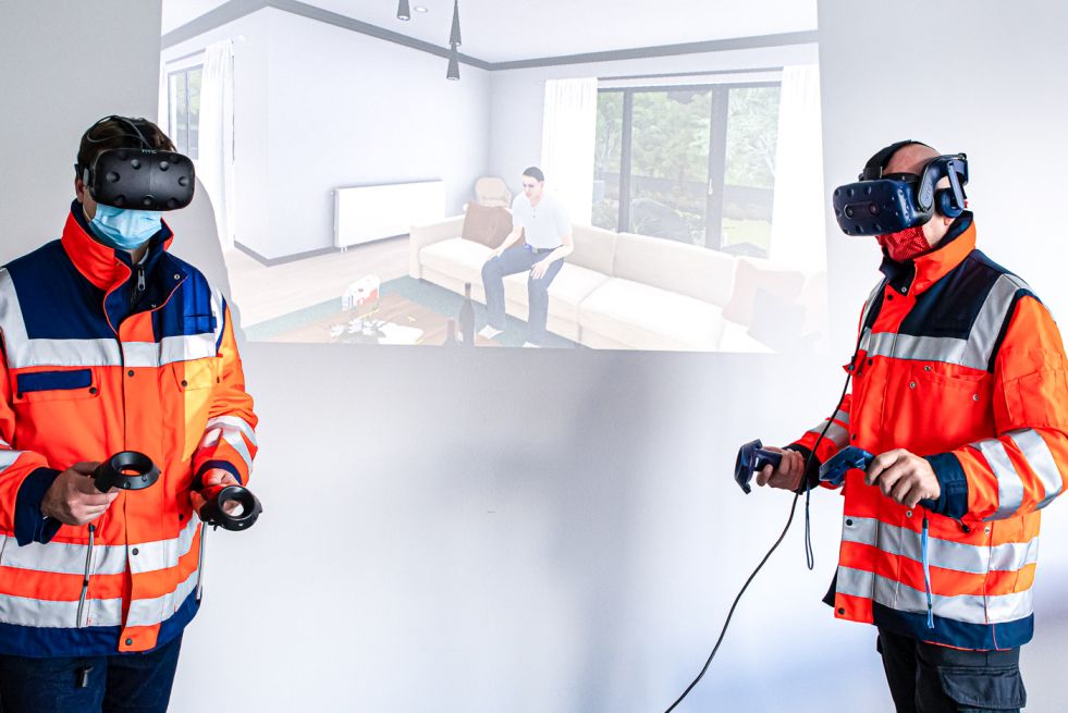 Auf dem Bild sind zwei Personen in DRK-Jacken zu sehen, die VR-Brillen tragen und deren virtuelle Erlebnisse mit einem Beamer für Zusehende an die Wand projeziert sind.