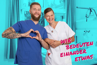 Auf dem Bild sind zwei Personen, wahrscheinlich Pflegekräfte, in DRK-Krankenhauskleidung zu sehen, die in die Kamera lachen. Eine Person formt mit ihren Händen ein Herz. Im Vordergrund steht "Wir bedeuten einander etwas".
