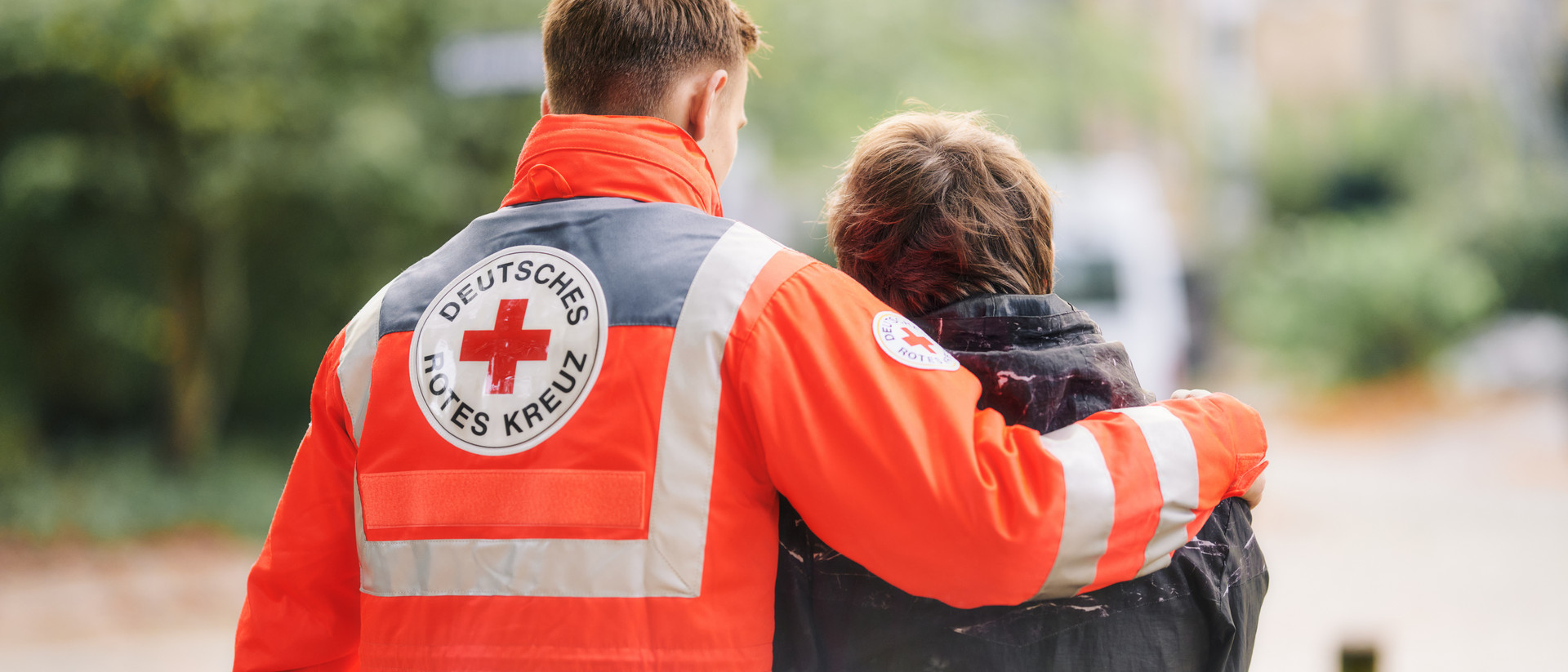 Umarmung Zwei Personen von hinten, eine trägt eine orangefarbene Jacke mit dem Logo des Deutschen Roten Kreuzes und legt den Arm um die andere Person