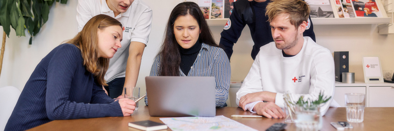 Eine Gruppe von Personen sitzt vor einem Laptop. Sie schauen sich gemeinsam etwas auf dem Gerät an und diskutieren.